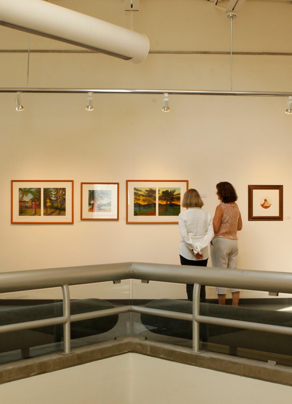 Two women look at a row of framed paintings hanging on a white wall in the Portland Campus art gallery